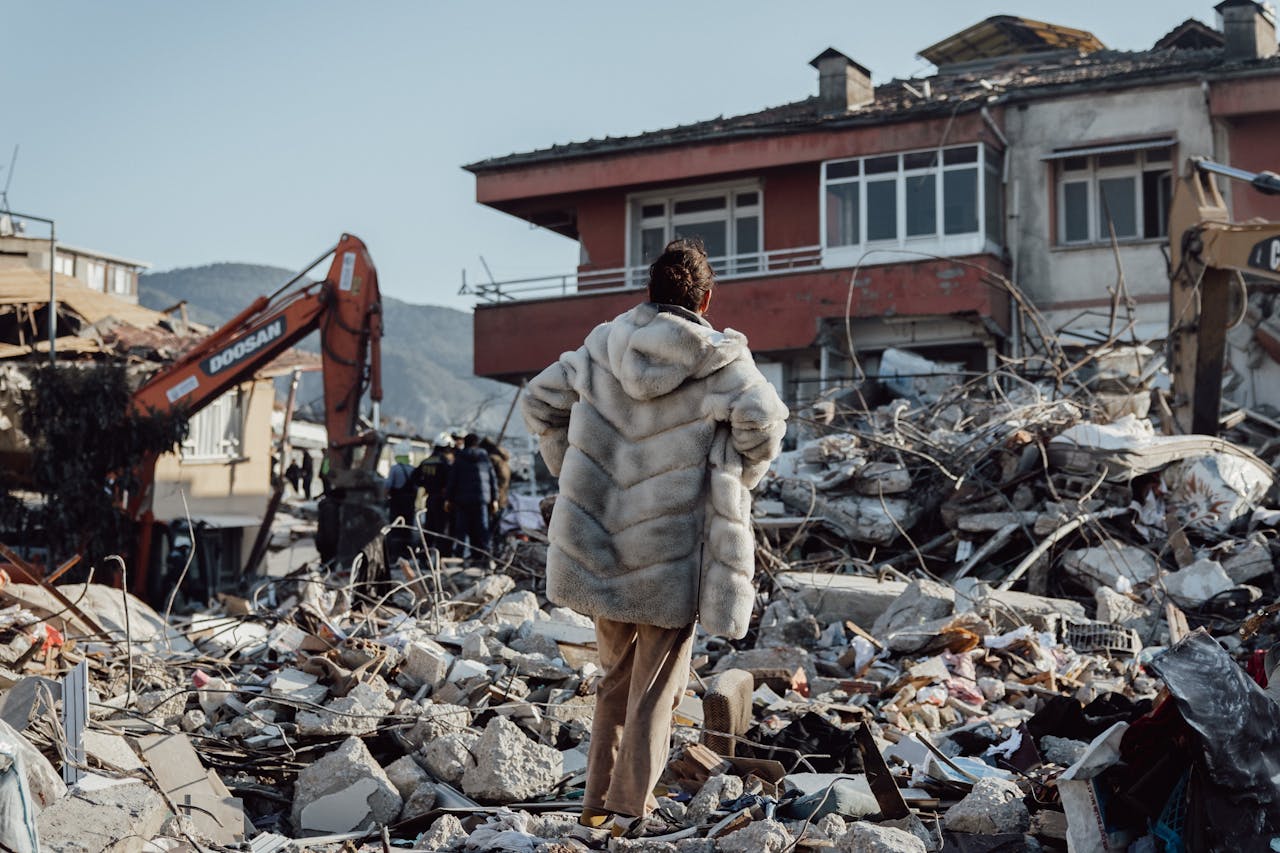 services-02 A woman in a fur coat stands amidst earthquake rubble, observing damaged buildings and an excavator.
